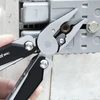 Pince d'un outil multifonction serrant une vis rouillée sur un boîtier gris électrique.
