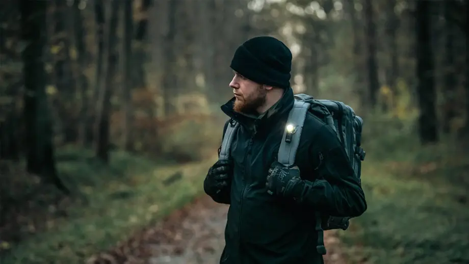 Randonneur barbu avec sac à dos et lampe, marchant sur un sentier dans la forêt.