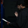 Homme avec une torche et un presse-papiers vérifiant les stocks dans un entrepôt sombre, manutention de nuit, inventaire.